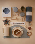 Flat lay of neutral-toned baby essentials, including a mushie Multi-Use Cover, cup, bowl with spoon, teether, stacking cups, pacifiers, cookie cutters, bib, utensils, and a rolled cloth—all arranged on a cream background.