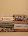 A neatly stacked pile of mushie Multi-Use Cover and folded clothes in soft neutrals and floral patterns sits against a beige background.