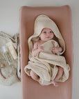 A baby wrapped in a beige towel lies on a soft pink changing pad. Beside the baby are a striped cloth, a bottle of baby oil, a tube of cream, and a pacifier clip with a wooden bead.
