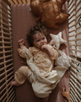 A curly-haired baby in pale yellow pajamas lies in a wooden crib, holding a pacifier and cuddling the mushie Star Lovey Blanket, with plush toys and a giraffe toy nearby.