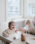 A smiling baby in a high chair is fed by a woman holding a small white dog. On the tray, a mushie Silicone Placemat made of food-grade silicone adds style and safety to the bright, naturally lit room.