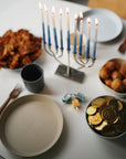 A Hanukkah table set with a lit menorah, latkes, sufganiyot, gelt, dreidels, utensils, empty dishes, and colorful mushie Dinnerware Cups—non-toxic and perfect for kids.