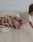 A baby in a cream outfit crawls on a light wooden floor next to a mushie Organic Cotton Muslin Swaddle Blanket, a pacifier, and a toy paint palette with colorful circles.