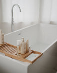 A modern white bathtub filled with water features a wooden bath tray holding skincare products, lotion, and mushie Baby Oil. Soft natural light filters through sheer curtains in the background.
