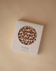 A boxed mushie Multi-Use Cover, shown against a beige background, features a leopard print and is ideal for use as a nursing or car seat cover.