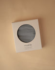 A boxed mushie Multi-Use Cover with a circular window displays the grey fabric inside, ideal for use as a nursing or carseat cover. Shown on a beige background.