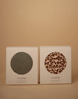 Two white boxes with round windows display mushie Multi-Use Covers—one in solid gray, the other in a brown and black leopard print. These versatile covers can be used for nursing or as a carseat cover.
