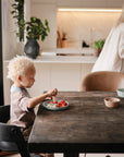 A young child with curly blonde hair sits at a wooden table eating fruit from mushie's Square Dinnerware Bowls, while an adult stands in the background of a modern kitchen. A glass of juice and two baby bowls are on the table.