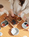 A woman cuts fruit on a wooden board while two young children sit at a table with mushie Lunchboxes featuring built-in compartments. Divided plates hold strawberries and blueberries, with extra berries and a wooden bowl on the table.