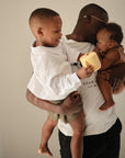 A man in glasses holds a smiling boy and a baby. The boy, in a white shirt, offers the mushie Snack Cup to the baby dressed in brown. The warm, joyful scene is set against a neutral background.