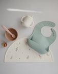 The mushie Silicone Placemat features floral designs on a light background and pairs perfectly with pastel-colored baby feeding items like a pale green bib, beige sippy cup with handles, and light pink bowl with matching spoon.