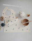 A flat lay of beige food-grade silicone baby feeding items on a white surface, featuring a whale print bib, mushie Silicone Placemat, bowl with spoon, sippy cup, pacifier, and stackable toys.