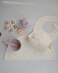A set of neutral and pastel baby feeding items—including a food-grade silicone bib, bowl with spoon, sippy cup, pacifier, and flower toy—all arranged on matching mushie Silicone Placemats for a stylish mealtime.