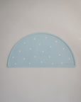 A light blue, semi-circular Silicone Placemat by mushie, made from food-grade silicone with small white daisy patterns and the embossed brand name in the lower right corner, is displayed on a plain, light-colored surface.