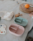 A table displays a mushie Lunchbox in pink, a mushie Lunchbox in green, each with built-in compartments, a silicone bib, a pop-it fidget toy, a wooden cutting board holding an orange and knife, and hands resting on the table.