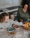 A woman slices oranges on a wooden board while two young girls reach for food at a kitchen counter filled with snacks, pastel plates, and a mushie Lunchbox featuring built-in compartments and made from non-toxic plastic.