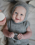 A smiling baby in a gray knit outfit with bunny ears lies on a soft surface, holding the mushie Koala Teether. An adult hand holds a cream container, while small dabs of cream are on the baby's cheeks.