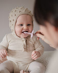 A baby in a beige knit outfit smiles as an adult helps brush their teeth with the pink mushie Flower Training Toothbrush.