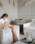 A woman in a white dress breastfeeds her baby wrapped in a mushie Organic Cotton Muslin Swaddle Blanket, sitting on a white armchair in a modern living room with neutral decor, pampas grass, and a nearby white sofa.