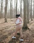 In a sunlit forest with tall, leafless trees and a ground of fallen leaves, a person in a white top and light pink pants holds their baby close using the mushie Baby Wrap.