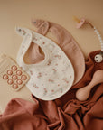 A flat lay of baby items on a beige background featuring mushie Muslin Bibs (one floral white, one solid pink), a teething toy, a silicone pop-it toy, and a soft rust-colored cloth.