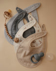 Four mushie Muslin Bibs in neutral tones, a pacifier clip with wooden beads, a silicone teether, and a clear pacifier are arranged neatly on a beige surface.