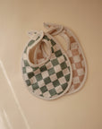 Two mushie Muslin Bibs made from organic cotton muslin, featuring large checkered patterns in green/white and brown/white, are displayed flat on a beige surface under soft lighting.