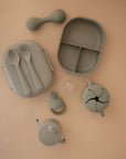 The mushie Snack Cup, in light green silicone, sits on a beige surface alongside matching baby feeding items: divided plate, lidded container with spoon and fork, sippy cup, pacifier, and teether—ideal for toddlers.