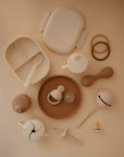 A flat lay of beige and brown baby items, including the mushie Lunchbox with built-in compartments, plates, cups, a spoon, teether, pacifier, sippy cup, beaded bracelets, and silicone toys arranged on a light brown background.