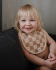 A smiling blonde-haired child sits on a dark couch, wearing a beige and brown checkered Muslin Bib by mushie. The organic cotton bib adds comfort, while the child looks content with hands resting in their lap.