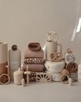 A neatly arranged display of mushie Lunchbox products in neutral tones, featuring non-toxic plastic containers, soft towels, pacifiers, teething rings, bottles, and folded blankets in beige, tan, and brown shades.