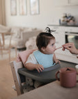 A baby with a topknot sits in a highchair, wearing a blue bib and opening their mouth for a spoonful from an adult. A mushie Snack Cup, designed to be spill-proof for toddlers, rests on the tray in the cozy, modern kitchen and living room.
