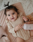A smiling baby with dark hair and a small ponytail lies on a pink quilted mat in a beige outfit. The baby is holding mushie Lavender Baby Body Lotion, while an adult hand helps apply it to their cheeks.
