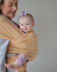 A smiling baby with a purple bow is carried in the mushie Baby Wrap on an adult’s chest. The breathable wrap keeps the baby close, while the adult looks down affectionately against a plain backdrop.