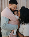 A man holds a baby in a pink mushie Baby Wrap made from organic cotton, while a woman gently kisses the baby's cheek. All three stand close together before a wooden wall, capturing the joy of baby wearing.