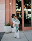 A woman in white clothes and grey sneakers stands on a sidewalk by a modern building, cradling her baby in a mushie Baby Wrap made from breathable organic cotton. Two large potted plants are nearby as her baby rests peacefully against her chest.