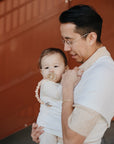A person in glasses holds a baby in a front carrier. The baby, dressed in neutral tones, uses a FRIGG Moon Silicone Pacifier 2-Pack by FRIGG. They stand before a rust-colored background—classic baby essentials.