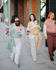 Three women stroll near modern buildings, smiling and holding coffee cups. Each wears casual clothes and carries a baby in a mushie Baby Wrap, designed for breathability and comfort during outings.