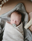 A baby sleeps peacefully in a crib, wrapped in a mushie Organic Cotton Muslin Swaddle Blanket with a pacifier. Nearby are a gray towel, a beige cream tube, and a soft pink cloth.