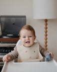 A smiling baby sits in a high chair wearing a patterned bib and holding the Mushie Silicone Fresh Food Feeder—perfect for encouraging safe self-feeding, with a lamp, plant, and record player visible on the shelf in the background.