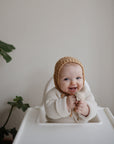 A smiling baby in a knitted brown bonnet and cream sweater sits in a high chair, holding the Mushie Silicone Fresh Food Feeder. Two green potted plants are in the background against a plain wall.