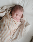 A newborn peacefully sleeps, wrapped in a mushie Organic Cotton Muslin Swaddle Blanket with beige and white stripes, lying on a soft cream-colored sheet.