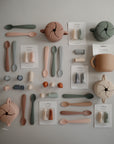 A flat lay of mushie silicone baby feeding essentials, featuring a spill-proof Snack Cup, training cups, soft spoons, finger toothbrushes, and anti-choke food feeders in neutral beige, sage green, blush pink, and gray.