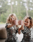 Two young girls in sparkly sequin dresses sit together, smiling and raising mushie Dinnerware Cups with decorative straws in a celebratory toast. Lush greenery is visible through the window behind them.