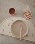 A curved, cream-colored silicone placemat with floral designs displays a matching round plate and pink fork. Nearby are mushie Dinnerware Cups in pink and a beige toy resembling a calculator, all set on a marble surface.
