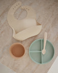 A mushie Silicone Feeding Spoon, a beige bib, a small tan bowl, and a green divided plate are arranged on a marble surface.