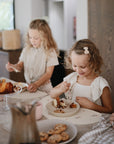 Two young girls sit at a kitchen table, smiling as they enjoy cake. Nearby are cookies and mushie Dinnerware Cups, while one girl holds a fork and the other focuses on her plate. A pitcher sits on the table too.