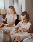 Two young girls sit at a bright, cozy kitchen table, smiling and eating cake on mushie Silicone Placemats. Plates of cookies and cake are laid out. One girl wears a hair clip and both are dressed in light-colored clothes.