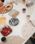 A white table is set for kids with mushie pastel, non-toxic plastic plates and cups, a mushie Dinnerware Fork and Spoon Set, plates of raspberries, blueberries, crackers, cereal bowls, and a glass water bottle. Kids gather around the table.