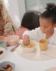 Two young girls sit at a table with bowls of food. One eats chips, while the other holds a stuffed animal. The table is set with pastel mushie Dinnerware Cups, matching dishes, and a placemat.